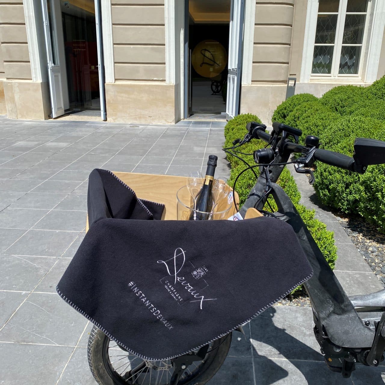 vélo dans une cour avec dans le panier seau à champagne, bouteille de champagne et flûtes et couverture de la marque Devaux